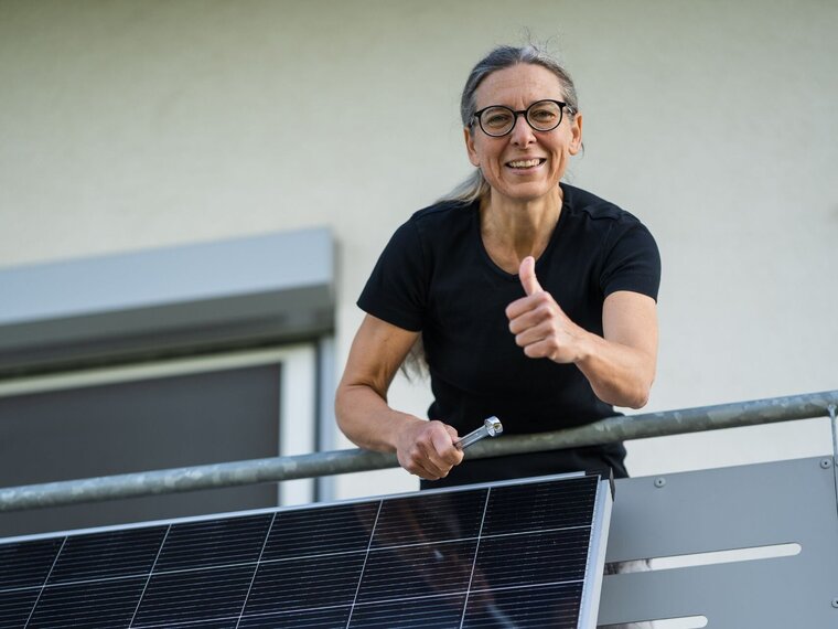 Eine Frau installiert ein Balkonkraftwerk auf ihrem Balkon. Sie lächelt und zeigt den Daumen hoch. 