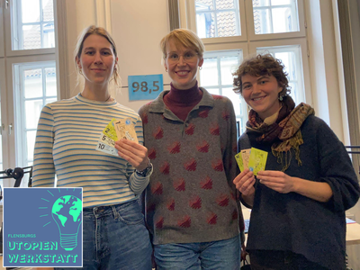 3 Menschen stehen nebeneinander und halten zeigen die bunten Klimabonus-Scheine in die Kamera. In der linken Ecke ist das Logo von Flensburg's Utopienwerkstatt zu sehen.